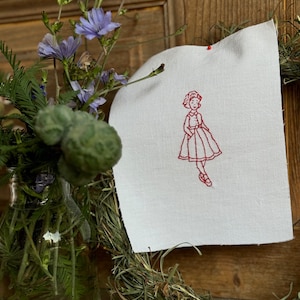 Könnte beinhalten: Ein weißer Stoff mit roter Stickerei einer Mädchen in einem Kleid. Der Stoff hängt an einer Holztür mit einem Kranz aus getrocknetem Gras.