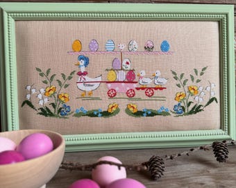 Könnte beinhalten: Eine gerahmte Kreuzstich-Stickerei, die eine skurrile Szene von Enten zeigt, die einen Wagen voller bunter Ostereier ziehen. Der Wagen ist mit Blumen verziert und die Enten tragen Hüte. Die Stickerei ist in einem grünen Holzrahmen gefasst.