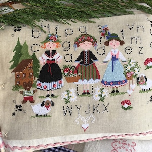 Puede incluir: Una almohada beige con un diseño de punto de cruz que presenta a tres chicas con vestidos tradicionales, una casa de jengibre, un oso y un perro. Las letras del alfabeto están bordadas alrededor del diseño.