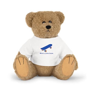 May include: A brown teddy bear wearing a white t-shirt with a blue skateboard graphic and the text "Mom's Little Dude".