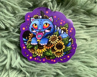 Op de afbeelding: Een sticker met een blauwe kat met oranje ogen, een kroon en een roze tong, omringd door zonnebloemen. De sticker heeft een paarse rand met gele stippen, op een groene, fuzzy achtergrond.