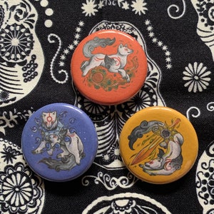 May include: Three round pinback buttons with illustrations of a white wolf with red markings. The buttons are orange, blue, and yellow. The background is a black and white patterned fabric.