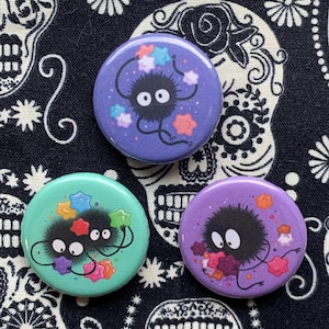 May include: Three round buttons with cartoon characters. One button is purple with a black character and colorful stars. Another is teal with two black characters and colorful stars. The last button is purple with a black character and colorful stars.