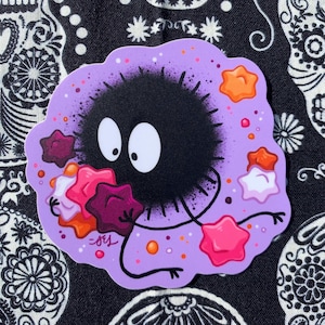 Puede incluir: Una pegatina con un personaje de dibujos animados, un duende de hollín, con cuerpo negro, ojos blancos y contorno morado. El duende está rodeado de objetos con forma de estrella en tonos rosa, naranja y morado. El fondo es blanco y negro.