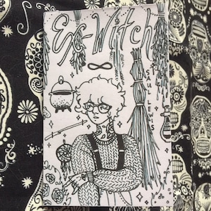 Peut inclure: Illustration en noir et blanc d'une personne aux cheveux courts bouclés portant un pull, une salopette et des lunettes. La personne tient un attrape-rêves et une baguette. L'image est entourée de divers symboles de sorcellerie et du texte "Ex-Witch" et "Saul Lewis".