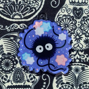 May include: A round, blue charm featuring a black, fuzzy creature with large white eyes and small arms and legs. The charm has a scalloped edge and is decorated with pastel-colored stars and dots. The background is a black and white patterned fabric.