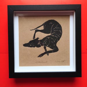 May include: A framed black and white linocut print featuring a dog in a dynamic pose. The dog's body is curved, and its legs are extended. The print is on tan paper, set within a black frame. The print is numbered 1/150.