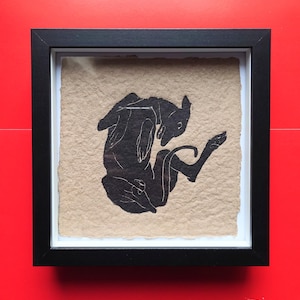 May include: Black silhouette art print of a dog in a black frame. The dog is depicted in a playful pose on a textured, tan background. The frame is square and has a white border. The background is a solid red.