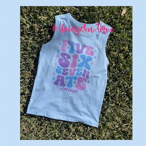 Pode incluir: Camisola sem mangas azul claro com as palavras "FIVE SIX SEVEN ATE" em rosa e roxo com um design de raio. O texto "CHEERLIFE" está impresso abaixo das palavras.
