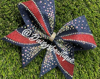 May include: A black and red cheer bow with rhinestones. The bow has a red stripe with a black background and is decorated with clear rhinestones. The bow is tied with a silver ribbon.