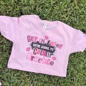 Pode incluir: Camiseta rosa cortada com o texto "Get in Loser We're Going to Cheer Practice" em letras pretas e rosa com lábios rosa ao redor do texto.