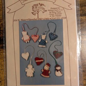 May include: A craft pattern packet titled Hickory Stick & Co. featuring illustrations of nine small fabric ornaments including hearts, a cat, an angel, a snowman, and a Santa figure, each with a hanging ribbon. The text includes address and contact details.