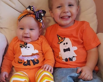 May include: Two children dressed in orange Halloween costumes. The older child is wearing a t-shirt with a ghost wearing a witch's hat. The younger child is wearing a onesie with a ghost wearing a tutu. Both children are smiling.