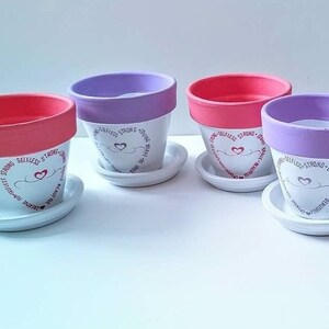 May include: Four white ceramic flower pots with pink and purple rims. Each pot has a white saucer and a pink heart with the words "Selfless, Strong, Loving, Brave" written around it.
