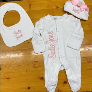 May include: A white baby outfit set featuring a bib, footed onesie, and a hat. The name "Sadie Jane" is embroidered in pink script on each item. The onesie has a ruffled edge and long sleeves. The hat is adorned with a pink bow.