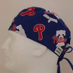 May include: Blue scrub hat featuring the Philadelphia Phillies logo in red and white. The hat has a tie closure and a small tag that reads "Unrattled Scrub Hats". The hat is designed to be worn during medical procedures.