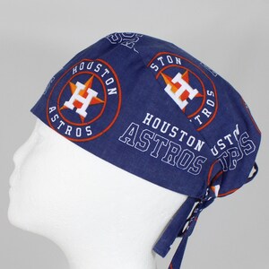 May include: Blue and orange surgical scrub hat with the Houston Astros logo repeated on the fabric. The hat has ties to tie it in the back.