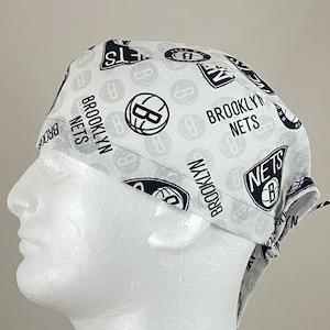 May include: White surgical scrub hat with a black and white pattern featuring the Brooklyn Nets logo and text. The hat is tied on a white mannequin head.