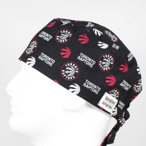 May include: Black scrub hat with a white and red Toronto Raptors logo pattern. The hat has a tie at the back.