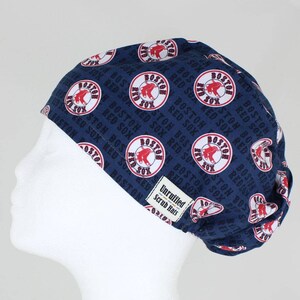 May include: A navy blue scrub hat featuring a repeating pattern of the Boston Red Sox logo and text. The logo is a red sock inside a white circle with the words "BOSTON RED SOX" around it. The hat has a tag that says "Unruffled Scrub Hats".