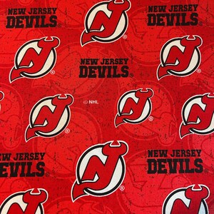 May include: Red fabric featuring the New Jersey Devils logo and team name in black and white. The logo is a stylized 'N' with a devil's horn, set within a white circle. The words "NEW JERSEY DEVILS" are printed in a bold font.