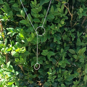 Puede incluir: Un collar de plata con dos colgantes circulares que cuelgan de una cadena. El collar se muestra sobre un fondo de follaje verde.