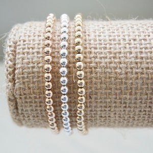 May include: Three beaded bracelets in gold, silver, and rose gold. The bracelets are stacked on a hessian background.
