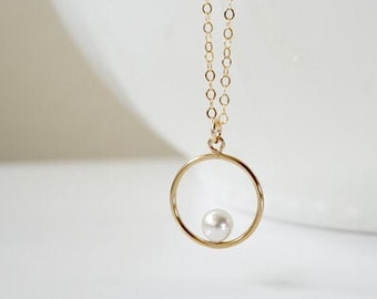 May include: A gold-toned necklace featuring a delicate chain and a circular pendant. A single, small, round white pearl is suspended in the center of the open gold circle. The metal has a polished finish, and the chain consists of small, uniform links.