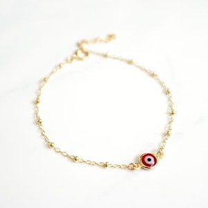 May include: A gold chain bracelet with a small red evil eye charm. The chain is made of tiny gold beads.