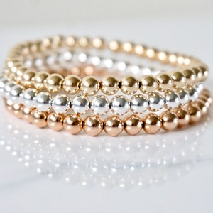 May include: Four beaded bracelets in gold, silver, and rose gold tones are stacked on a white surface. The bracelets are made of small, round beads, reflecting light. The bracelets are a jewelry item.