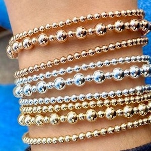 May include: A stack of gold, silver, and rose gold beaded bracelets. The bracelets are layered on a person's wrist.