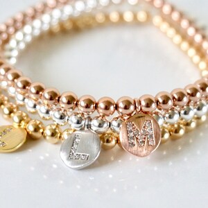 May include: Three beaded bracelets in gold, silver, and rose gold. Each bracelet has a different color of round beads. The bracelets have round, flat charms with the letters "L" and "M" encrusted with small, clear crystals.