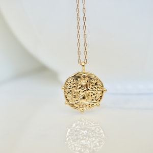 May include: Gold chain necklace with a round pendant featuring an intricate design of a person standing on a platform. The pendant has a textured surface and is surrounded by small decorative accents.