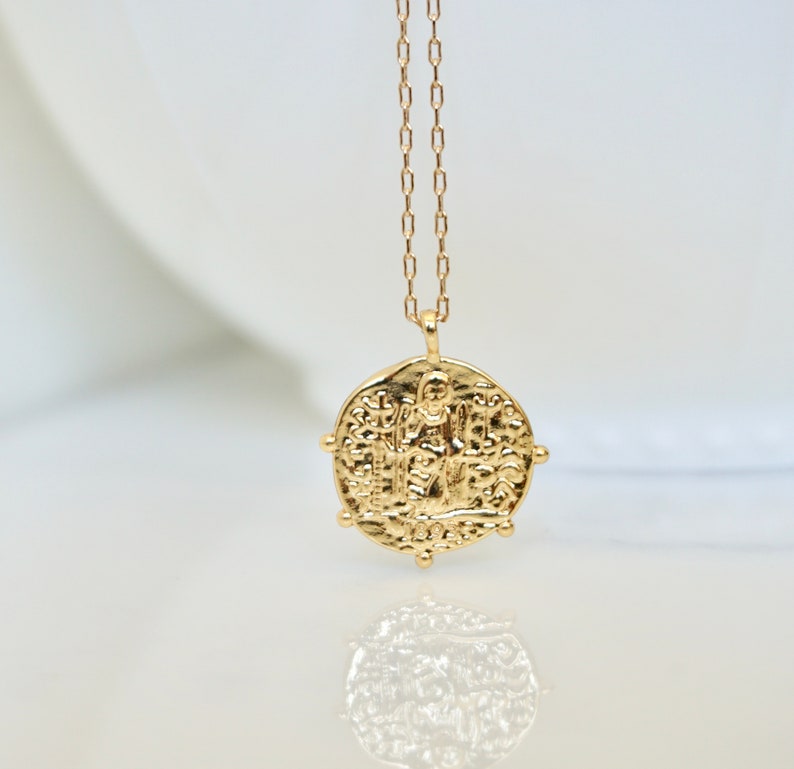 May include: Gold chain necklace with a round pendant featuring an intricate design of a person standing on a platform. The pendant has a textured surface and is surrounded by small decorative accents.