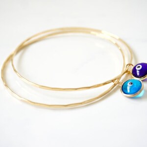 May include: Two gold-colored bangles with a hammered texture. Each bangle has a charm with an evil eye design. One charm is blue and the other is purple, both with gold-colored settings.