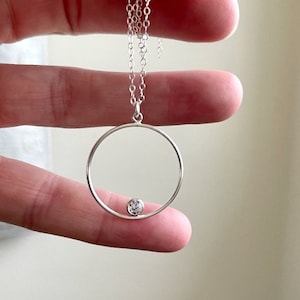 Puede incluir: Un collar de plata con un colgante circular y una pequeña piedra transparente en el centro.