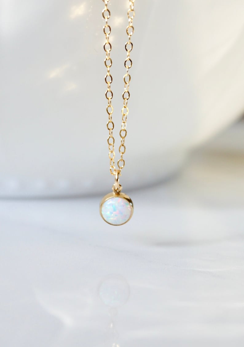 May include: A delicate gold chain necklace with a small, round opal pendant. The opal is white with a hint of iridescent color.