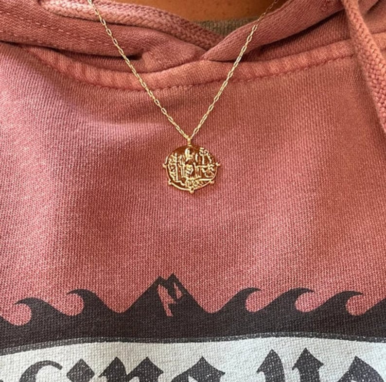 May include: A gold chain necklace with a round pendant featuring an intricate design. The pendant is worn on a pink hoodie with a black and white wave design.