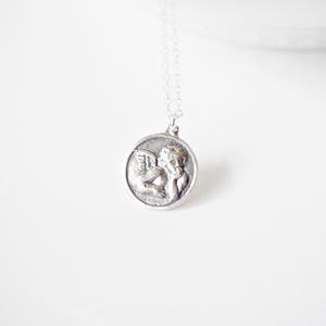 Puede incluir: Collar de plata con un colgante redondo que representa un querub&iacute;n alado.