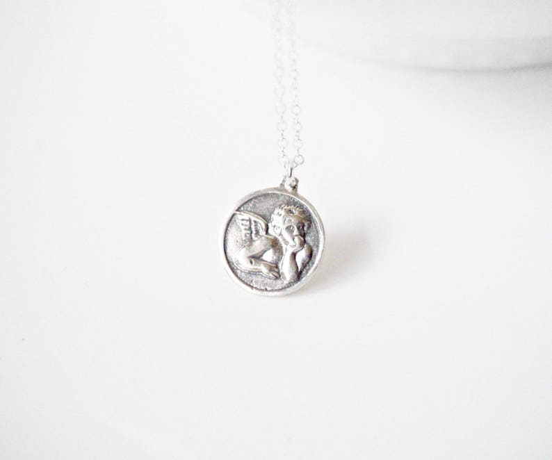 Puede incluir: Collar de plata con un colgante redondo que representa un querub&iacute;n alado.