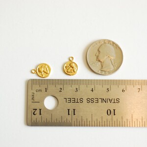 Puede incluir: Dos abalorios de metal de color dorado con un dise&ntilde;o de dos figuras que se toman de la mano. Los abalorios se muestran junto a una regla y una moneda de 25 centavos de d&oacute;lar estadounidense para comparar el tama&ntilde;o.