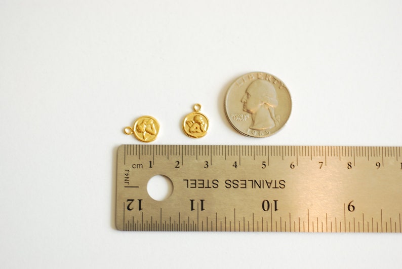 Puede incluir: Dos abalorios de metal de color dorado con un dise&ntilde;o de dos figuras que se toman de la mano. Los abalorios se muestran junto a una regla y una moneda de 25 centavos de d&oacute;lar estadounidense para comparar el tama&ntilde;o.