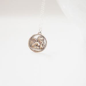 Puede incluir: Collar de cadena de plata con un colgante redondo que presenta una escultura en relieve de un &aacute;ngel con alas.
