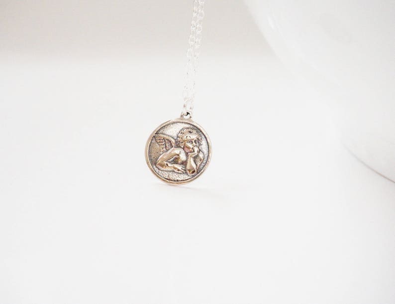 Puede incluir: Collar de cadena de plata con un colgante redondo que presenta una escultura en relieve de un &aacute;ngel con alas.
