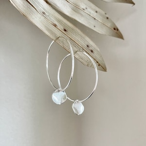 May include: A pair of silver hoop earrings with a small, round, white pendant attached to each earring. The earrings are displayed against a neutral background with a dried leaf.