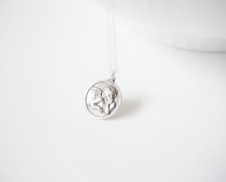 Puede incluir: Collar de plata con un colgante redondo que presenta una imagen detallada de un &aacute;ngel con alas.