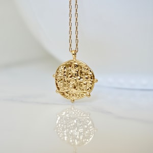 May include: A gold necklace with a round coin pendant featuring an intricate design. The coin is textured and has a raised edge.