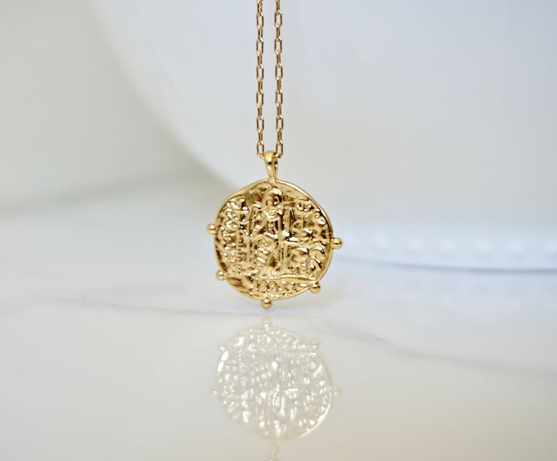 May include: A gold necklace with a round coin pendant featuring an intricate design. The coin is textured and has a raised edge.