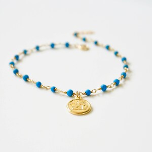 May include: A gold chain bracelet with blue beads and a gold charm. The charm is a round disc with a design on it.