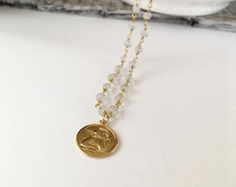 May include: A gold-toned necklace featuring a circular pendant with an embossed cherub figure. The chain is composed of small, translucent, faceted beads linked together with thin gold-colored wire, creating a delicate and refined appearance.